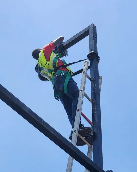 A worker in a safety harness at the top of a pole, against a blue sky